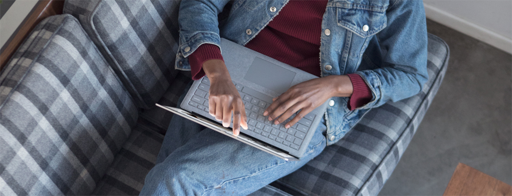 A Woman Wearing Denim Lying Back on the Couch Touching Her Microsoft Surface Laptop's Open Screen